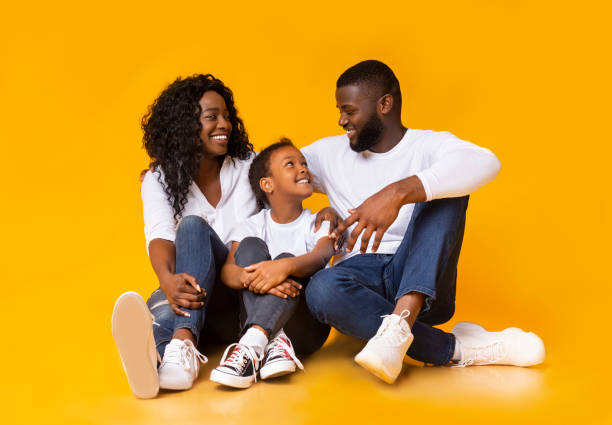 Black parents embracing with their adopred daughter, sitting on floor together Adoption concept. Happy afro parents bonding and embracing with their adopted daughter, sitting on floor over yellow background in studio yellow husband stock pictures, royalty-free photos & images