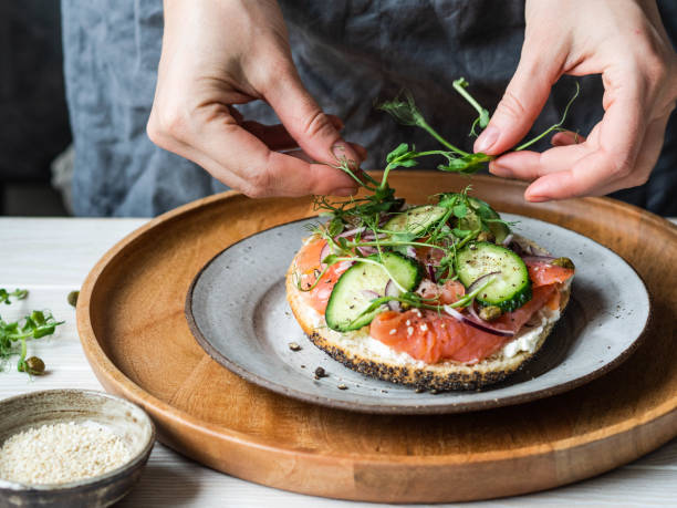 Healthy toast with cream cheese, salmon, fresh cucumber, capers, red onion, black pepper and and an uncertain woman lays on sandwich pea sprouts . stock photo