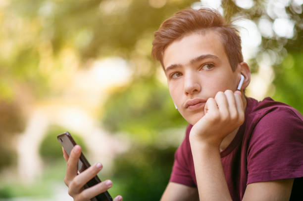 Close-up portrait of a thoughtful unhappy teenage boy with smartphone, outdoors. Sad teenager with mobile phone looks away, in the park. Pensive teenager in casual clothes with cell phone in park retrato de cerca de un adolescente reflexivo infeliz con teléfono inteligente, al aire libre. triste adolescente con teléfono móvil mira hacia otro lado, en el parque. adolescente pensativo en ropa casual con teléfono celular en el parque