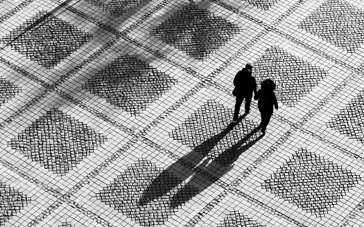Black and white toned image depicting an aerial high angle view of a couple (a man and a woman) walking and holding hands across a pedestrianised city square. The sun is low in the sky and the couple cast long shadows across the paved square behind them. Room for copy space.