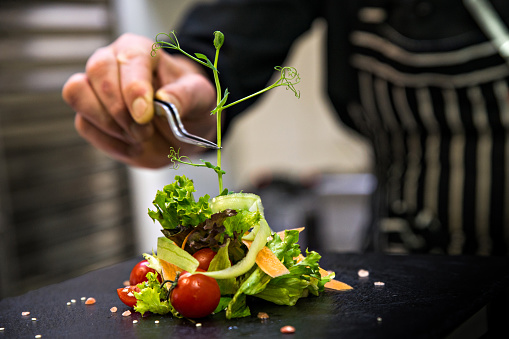 Chef finishing healthy salad on a black plate with tweezers. almost ready to serve it on a table.