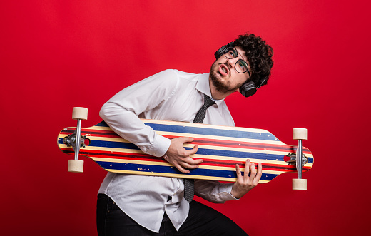 A young man with headphones and longboard in a studio on red background, having fun.