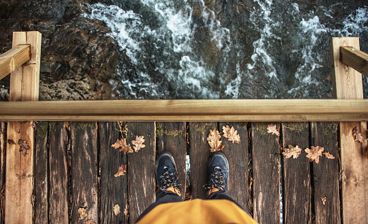 Directly above view of man with hiking boots standing on a wooden bridge crossing a water stream in autumn