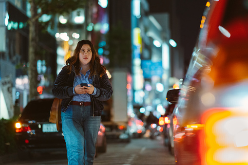 A young woman is holding a smart phone and looking for directions while walking in the city at night.