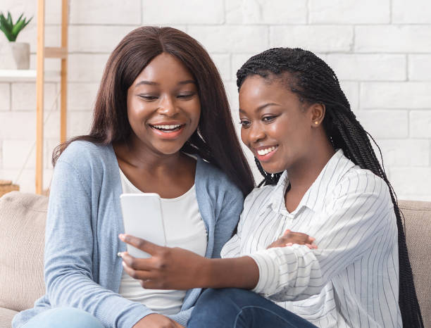 istock Two cheerful black girlfriends taking selfie, sitting on couch at home 1191897376