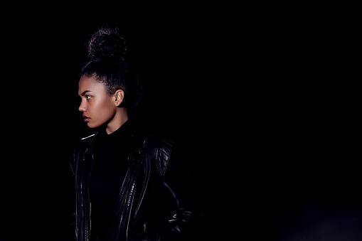 a young African American girl in a leather jacket and black clothes poses on an abandoned sand road. at night in the light of car headlights and lanterns