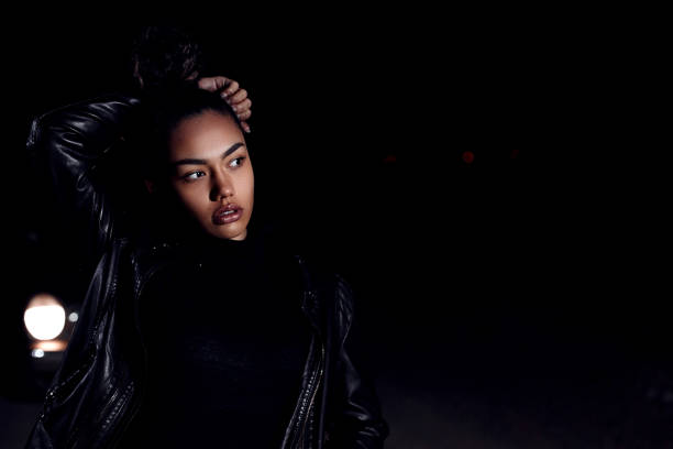 une jeune fille afro-américaine dans une veste en cuir et des vêtements noirs pose sur une route de sable abandonnée. la nuit à la lumière des phares de voiture et des lanternes - femme fatale one person make up cool photos et images de collection