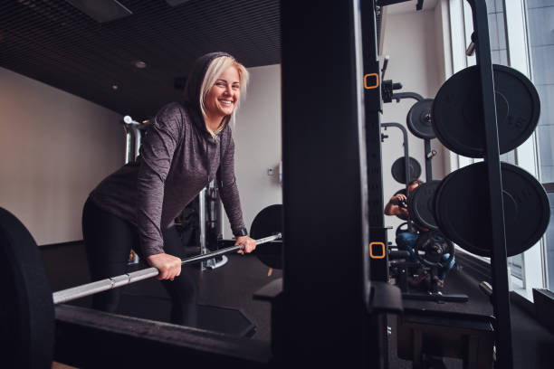 Sportive girl wearing hoodie leaning on a barbell next to a stand, smiling and looking at a camera Beautiful sportive girl wearing hoodie leaning on a barbell next to a stand, smiling and looking at a camera blonde female bodybuilders stock pictures, royalty-free photos & images