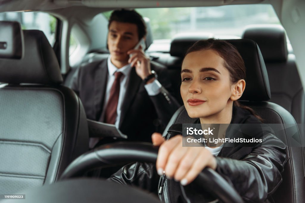 Woman taxi driver holding steering wheel while businessman talking on smartphone Taxi Driver Stock Photo