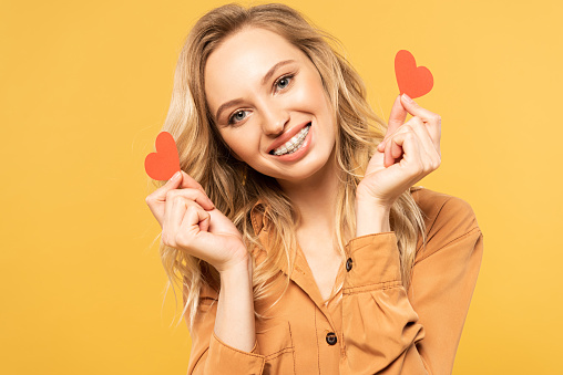 Smiling woman with dental braces holding paper hearts isolated on yellow