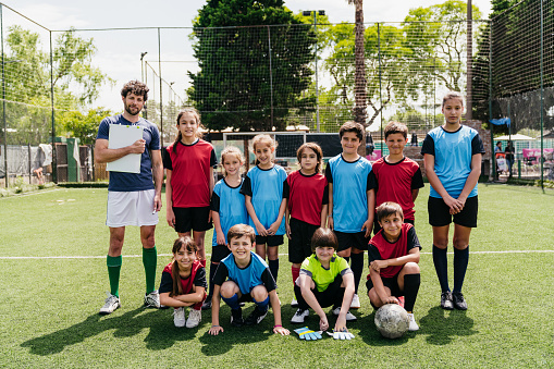 Two kids soccer teams with the coach posing on a soccer field. Red and blue uniforms. Looking at camera.