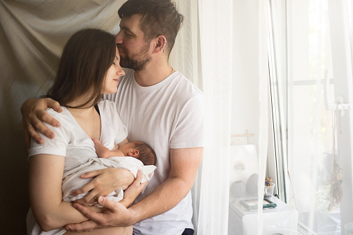 Father and mother with a newborn son standing by the window. New young multi-generation small family. Breast-feeding home concept