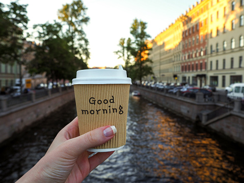Mockup of female hand holding a coffee paper takeaway cup on the river background with copy space. Close-up view of disposable paper cup of coffee in woman's hand. Woman holds a cup of morning coffee close up.