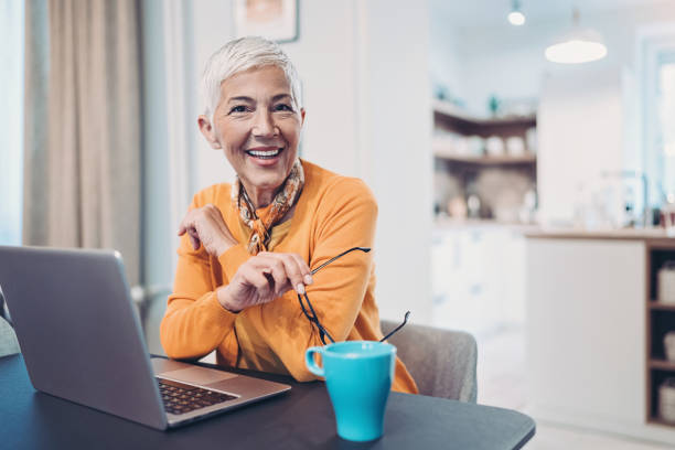 mulher sênior com um portátil e um café confortáveis em casa - looking at camera senior women laptop only women - fotografias e filmes do acervo