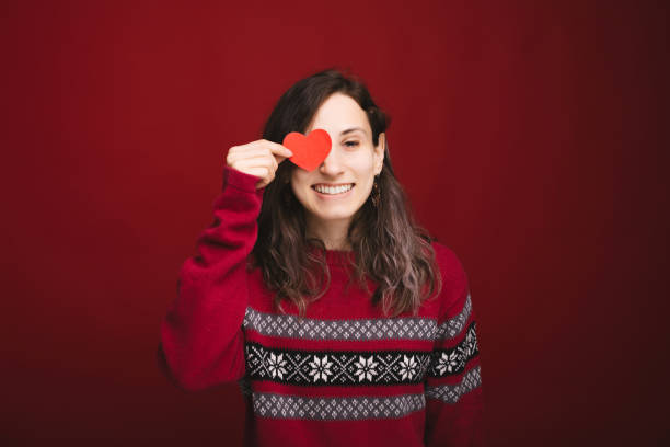 mulher nova encantadora no sweeater vermelho que prende o coração de papel vermelho sobre o olho - child making a face human face humor - fotografias e filmes do acervo