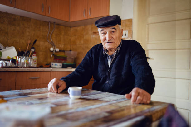 一人で座っている自宅のテーブルのそばにコーヒーや紅茶を飲みながら、黒いセーターと帽子をかぶったシニアマン祖父老農家 - one senior man only grandfather portrait old ストックフォトと画像