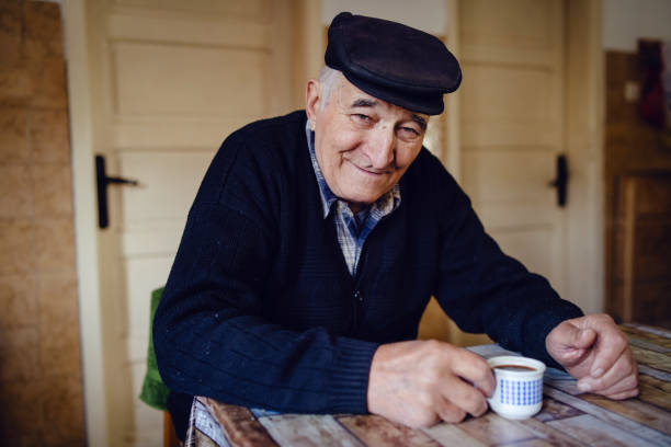 黒いセーターと帽子をかぶったシニアマン祖父の年金農家は、一人で笑顔で家のテーブルのそばにコーヒーや紅茶を飲む - one senior man only grandfather portrait old ストックフォトと画像