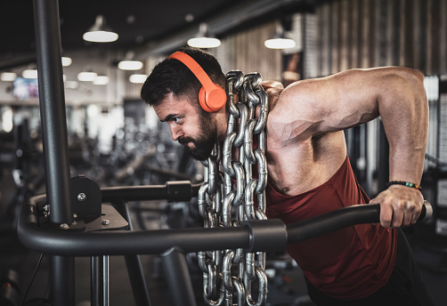 Side view of male strong bodybuilder training with weight steel chain, listening music with wireless headphones