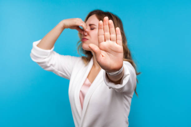 Bad smell. Portrait of woman pinching her nose and showing stop gesture, expressing disgust to unpleasant odor. blue background Bad smell. Portrait of woman with wavy hair in white jacket pinching her nose and showing stop gesture, expressing disgust to unpleasant odor, fart gases, her eyes closed with abhor. blue background unpleasant smell stock pictures, royalty-free photos & images