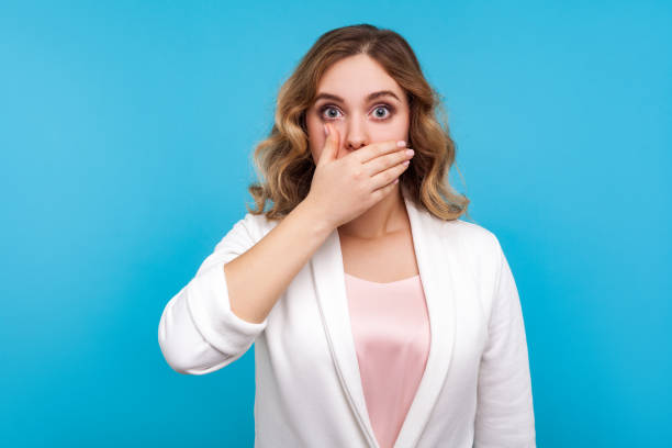 don't tell! portrait of frightened woman covering mouth with hand scared to speak. isolated, blue background - silence finger on lips businesswoman one person imagens e fotografias de stock