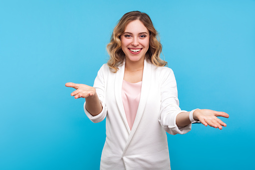 Please take! Portrait of kind cheerful woman with wavy hair in white jacket raised hands as if sharing, giving for free, offering hugs with friendly generous face. indoor studio shot, blue background