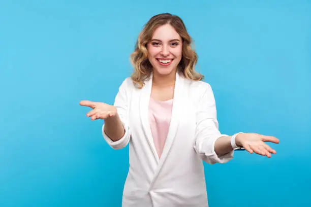 Please take! Portrait of kind cheerful woman with wavy hair in white jacket raised hands as if sharing, giving for free, offering hugs with friendly generous face. indoor studio shot, blue background