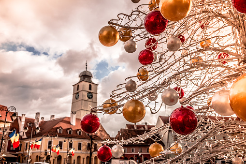 Color image depicting a metallic Christmas tree decorated with red and gold baubles in the medieval Transylvanian city of Sibiu, Romania. Beyond the tree we can see the beautiful traditional architecture and clock tower of the main square. Romanian flags hang from the street lamps, and the image is offset by a beautiful blue sky and cloudscape.