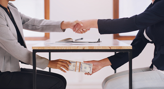 Cropped shot of an unrecognizable female financial advisor receiving a bribe under her table while shaking hands with her client in her office