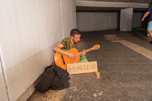 Photo of Homeless and Depressed Man who is Sitting in the Train Station, Subway Station, Tunnel or Underground and Playing Guitar with Homeless Sign and Donation Box. Desperate Mature Man is Feeling Abandoned and Lost in Depression and Sitting on Ground Street While Suffering Emotional Pain. Poverty and Social Issue Concept.