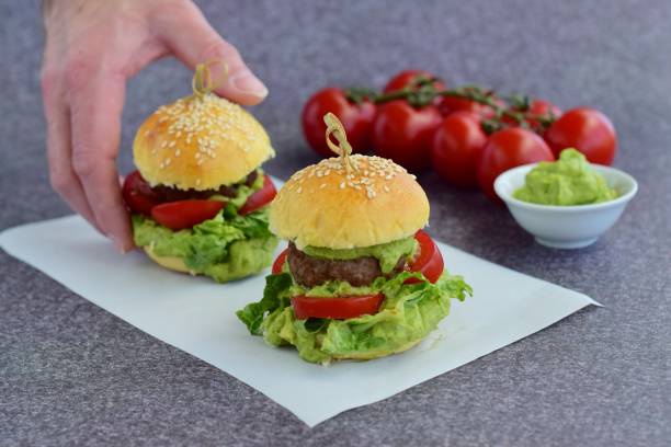 A hand picking up a burger. Sesame buns with beef patties, guacamole, tomato and lettuce