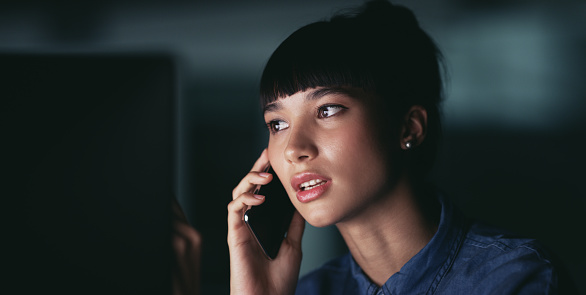 Shot of a young businesswoman talking on a cellphone while working on a computer in an office at night