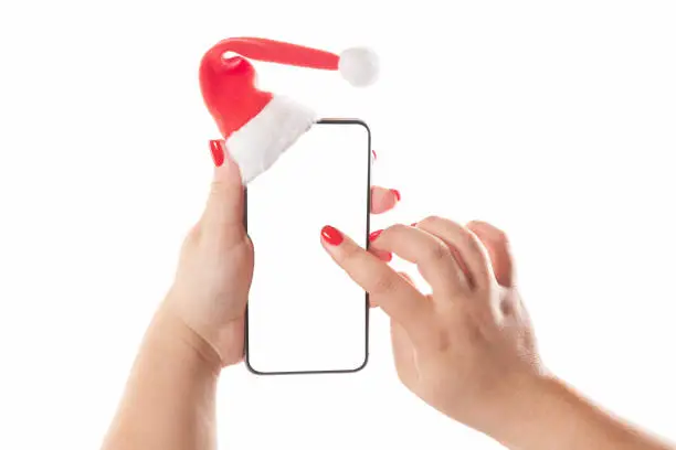 Female hands holding mobile smart phone with santa hat isolated on white background. Blank white screen. Concept of Christmas congratulation.