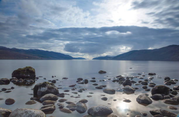 una vista a través del lago ness mirando por la longitud del lago con rocas posada el primer plano y nubes oscuras por encima, en escocia - loch ness fotografías e imágenes de stock