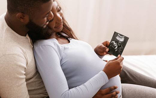 Relaxed african couple watching baby ultrasound photo, lying in bed at home, free space