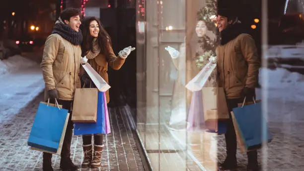 Xmas Seasonal Sales. Girl Pointing At Shopwindow Shopping With Boyfriend In Winter City In Evening. Panorama, Copy Space