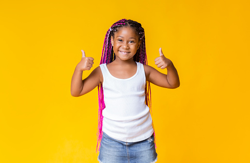 Like it. Pretty little african american girl gesturing thumbs up and smiling over yellow studio background with copy space