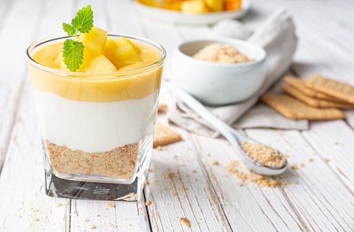 Sweet layered dessert with crushed graham crackers, cream cheese and pineapple curd, topped with fresh fruit slices in a glass jar on old wooden background