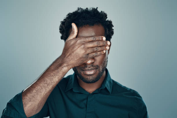 schöne junge afrikanische mann bedeckt augen mit der hand, während stehengrau hintergrund - hands covering eyes stock-fotos und bilder