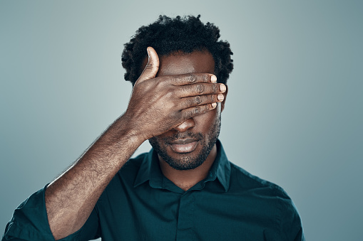 Handsome young African man covering eyes with hand while standing grey background