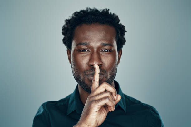 istock Handsome young African man looking at camera and keeping finger on lips standing against grey background 1192096274
