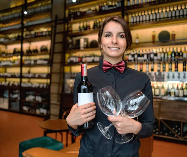 Female sommelier working at a wine bar holding glasses and a bottle of wine and looking at the camera smiling
