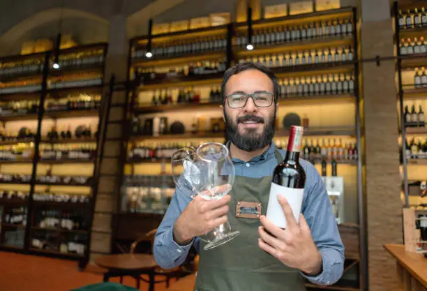 Happy sommelier working at a wine bar and holding a bottle with glasses and looking at the camera smiling