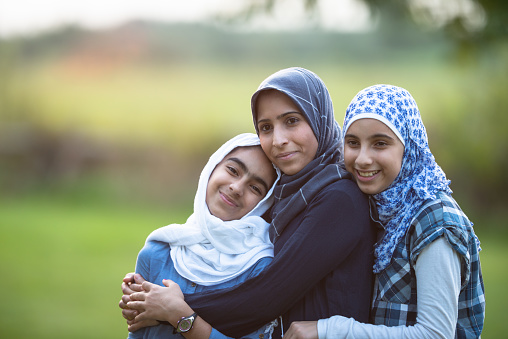 A young Muslim mother and her two daughters stand outside on a sunny summer day posing for a portrait.  They are all in close and hugging one another.  They are each dressed casually and wearing Hijabs.