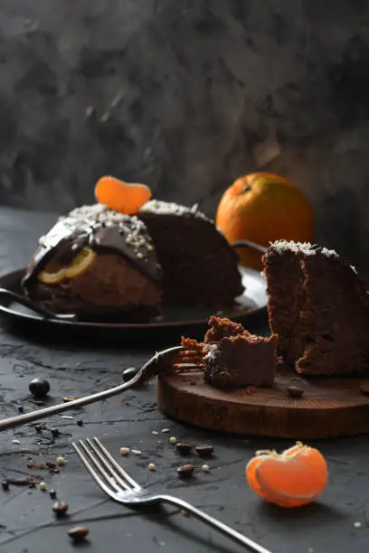 Homemade winter dessert. Traditional British pudding with oranges, chocolate icing and coconut shreds cut on wood slab on black background. Low key still life high angle view