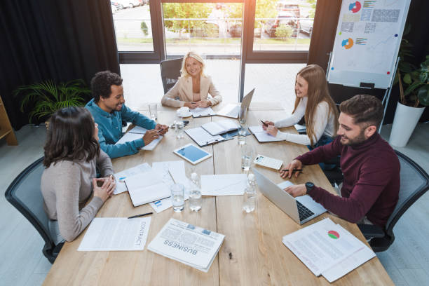 empresarios multiculturales positivos sentados en un escritorio de madera cerca de dispositivos digitales, documentos y vasos con agua en la sala de reuniones - office water business meeting fotografías e imágenes de stock