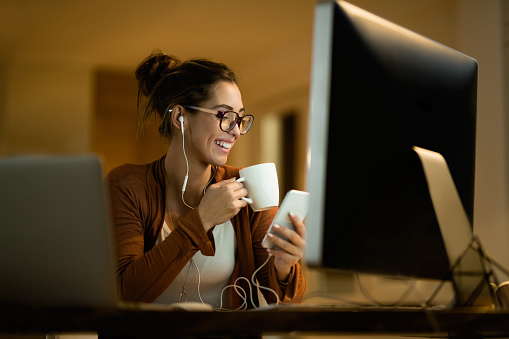 Young happy woman using cell phone while drinking tea and working at night at home.