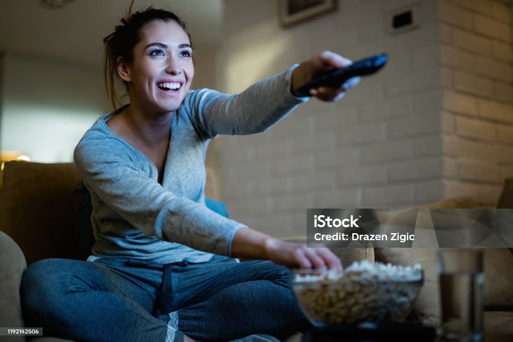 This is my favorite episode! Young happy woman changing channels with remote control while watching TV and eating popcorn in the evening at home. Watching TV Stock Photo
