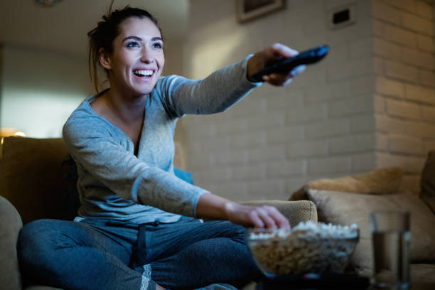 This is my favorite episode! Young happy woman changing channels with remote control while watching TV and eating popcorn in the evening at home. watching tv stock pictures, royalty-free photos & images