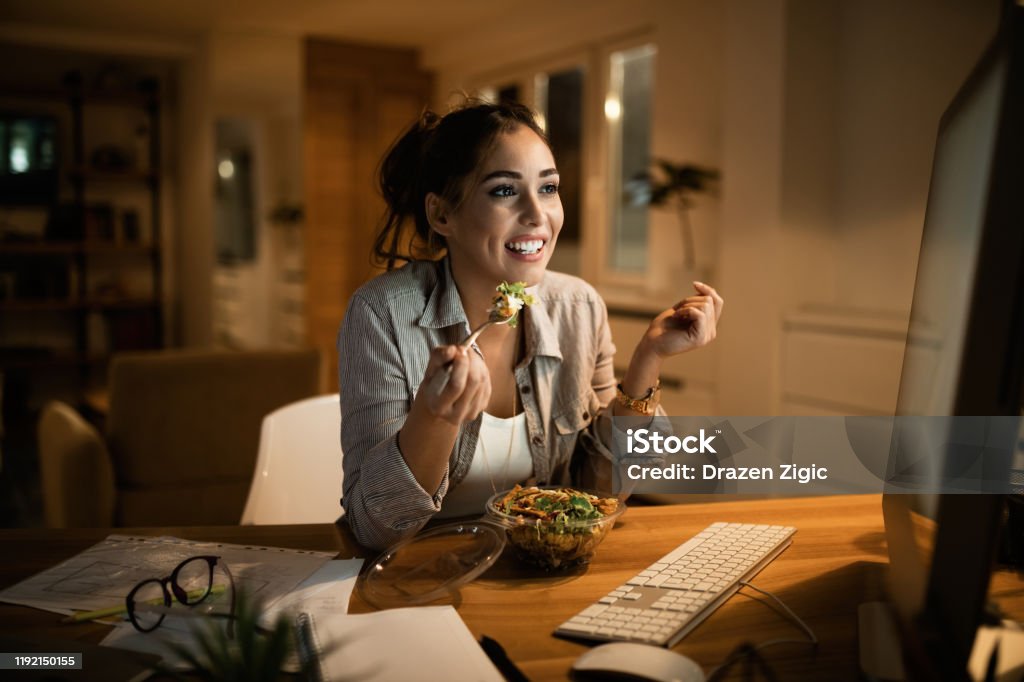 Young woman eating salad while working on a computer late at night. Smiling woman reading an e-mail on desktop PC and eating salad in the evening at home. Eating Stock Photo