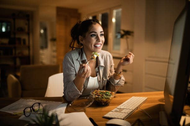 Young woman eating salad while working on a computer late at night. Smiling woman reading an e-mail on desktop PC and eating salad in the evening at home. evening meal stock pictures, royalty-free photos & images